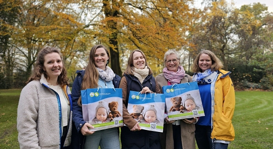 Fünf Frauen stehen auf einem grünen Rasen. Sie schauen in die Kamera und halten eine Tasche in der Hand. Auf der Tasche ist ein Foto von einem Baby mit einem Teddybär zu sehen.