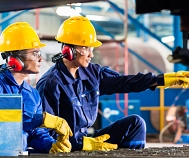 Ein Mann und eine Frau sitzen in blauer Arbeitskleidung und mit gelben Helm vor einer Maschine in einer Halle. © pixabay