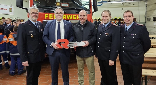 Fünf Männer, drei davon in Feuerwehruniform stehen vor einem roten Feuerwehrauto und halten einen großen roten Schlüssel in die Hand. Im Hintergrund stehen Mitglieder der Jugendfeuerwehr in neonfarbenen Jacken.